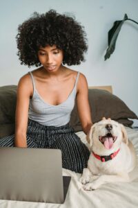 African American female surfing internet on netbook while stroking dog with tongue out on cozy bed