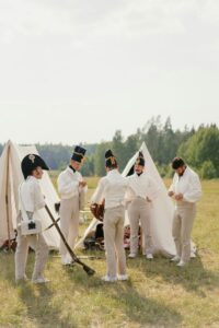 Men in French military uniform standing during reconstruction on grassy ground near tents and trees in countryside