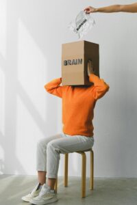 Crop anonymous person putting creased paper with Rubbish inscription in container with Brain title on head of female on chair