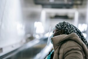 Black woman in face shield and mask standing near escalator